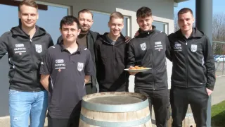 Six men in matching black jackets pose for a photo in front of a building with a wooden barrel. Each man holds a plate of food, and they are smiling.