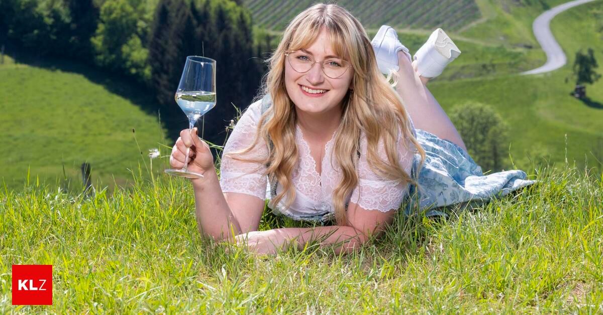 A woman with blonde hair and glasses lies on the grass, smiling and holding a wine glass. The background features a vineyard and a hill.