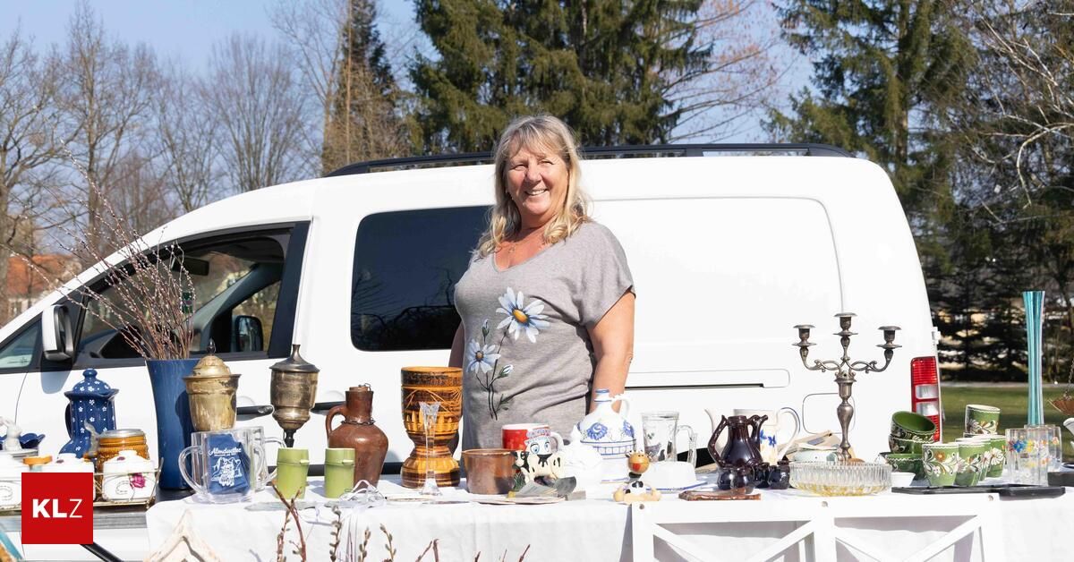 A woman with blonde hair is standing behind a table filled with various items. The table has a white tablecloth and several items, including a mug, a vase, and a candle holder. The woman is smiling and seems to be posing for a photo. Behind her, there is a white van, and there are trees in the background.