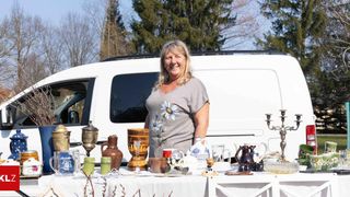 A woman with blonde hair is standing behind a table filled with various items. The table has a white tablecloth and several items, including a mug, a vase, and a candle holder. The woman is smiling and seems to be posing for a photo. Behind her, there is a white van, and there are trees in the background.