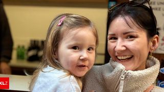 A woman and a young girl are smiling and looking at the camera. The girl has her hair in pigtails. They are standing close together.