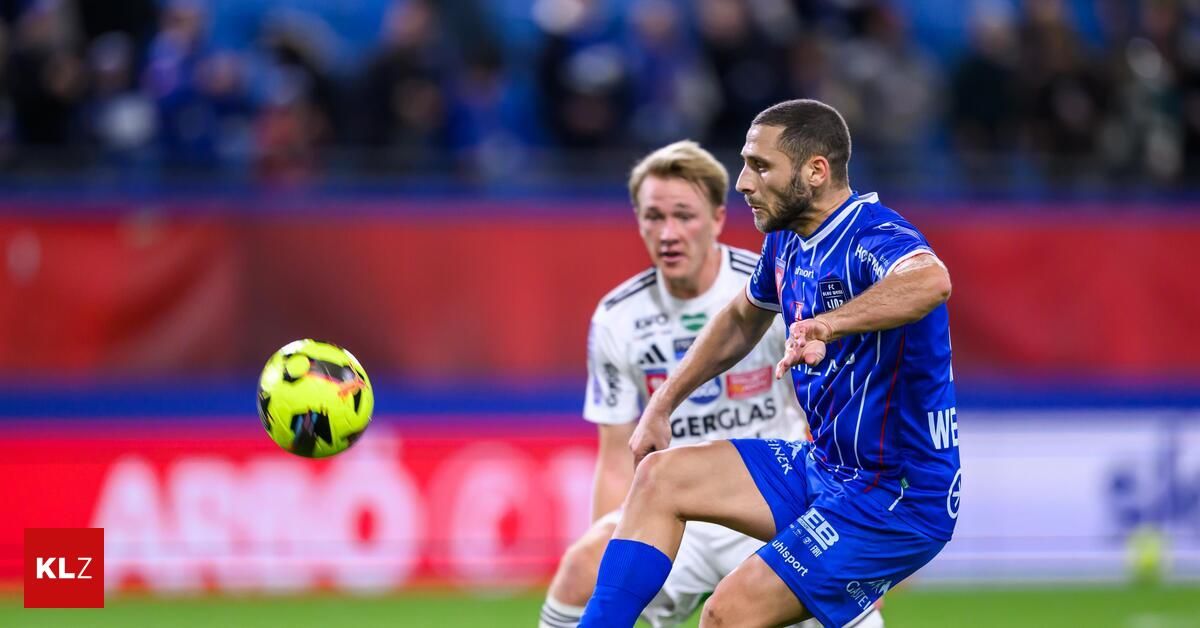 A soccer player in a blue jersey is kicking a ball in the air. Behind him, another player in a white jersey is looking at the ball. The ground has a red and white pattern.