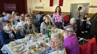A group of women sit at a table, smiling for a photo. They are enjoying a meal, with plates of food, drinks, and flowers on the table. Behind them, a woman stands and smiles, and a child stands near the door.