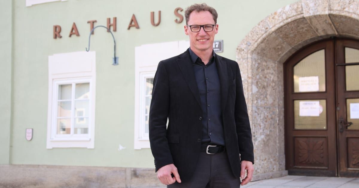 A man stands outside a building with the word 'Rathaus' on it, smiling. He is wearing a black blazer, a blue shirt, and glasses. The building has a lamp, windows, and a door.