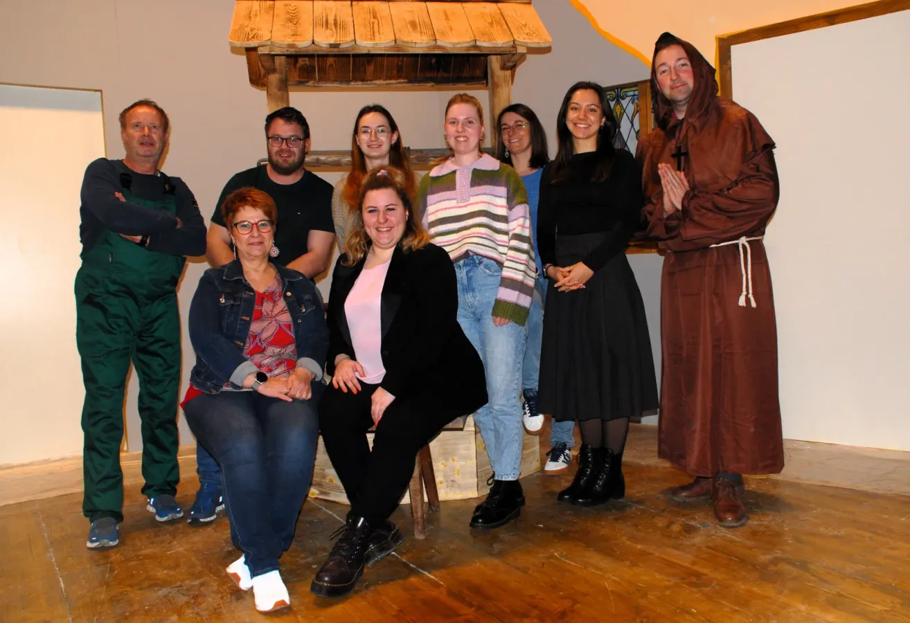 A group of people pose for a photo in a room. One person in a monk's robe stands on the right. A wooden structure is behind them.