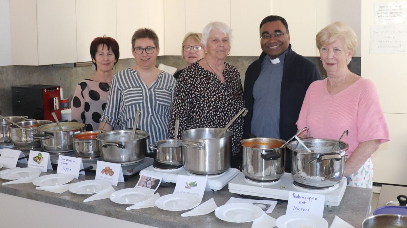 A group of people stand behind a counter with pots and plates. They are smiling. A sign reads 'Rohmsuppe mit Nocher!' and other food items.