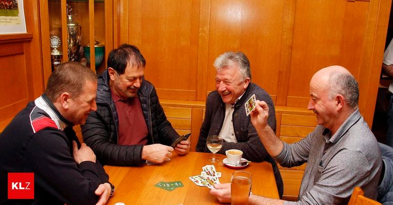 Vier Männer sitzen um einen Holztisch, lächeln und lachen, während sie ein Kartenspiel spielen. Jeder Mann hält eine Karte, und auf dem Tisch stehen Gläser und eine Untertasse mit Kaffee. Im Hintergrund befinden sich Holzwände und ein Schrank.