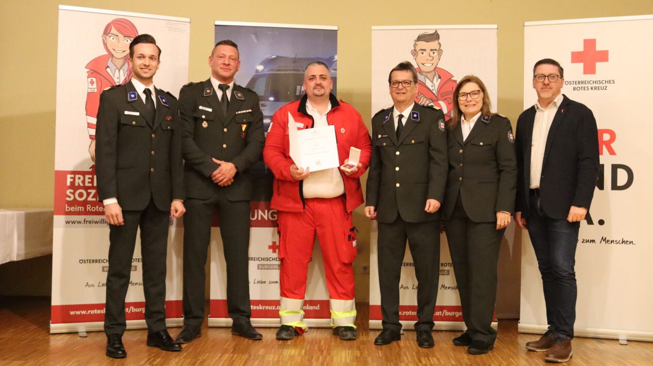Five individuals in uniforms stand together, one holding a certificate and a medal. Two on the sides are in military uniforms, and three in the middle, with one in a red suit. They are standing in front of banners.