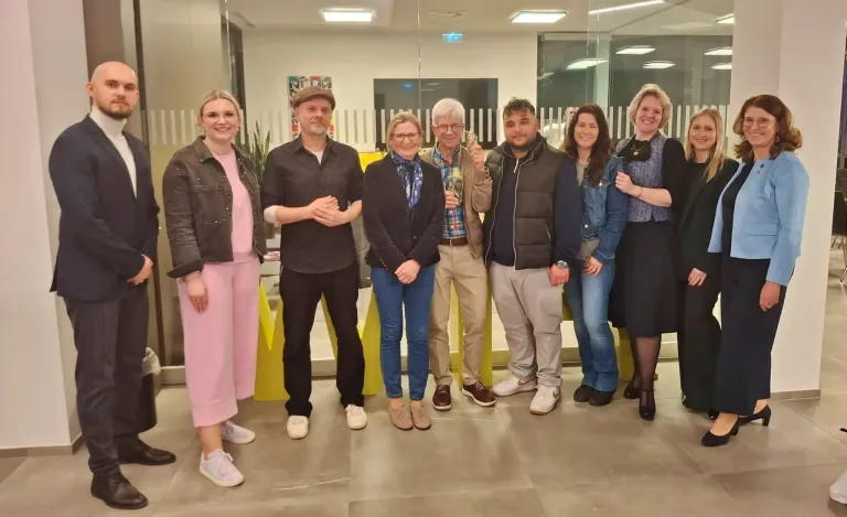 A group of people are posing for a photo in a modern office. The man in the middle holds a bottle. The people are dressed in various outfits, and there are yellow structures behind them.