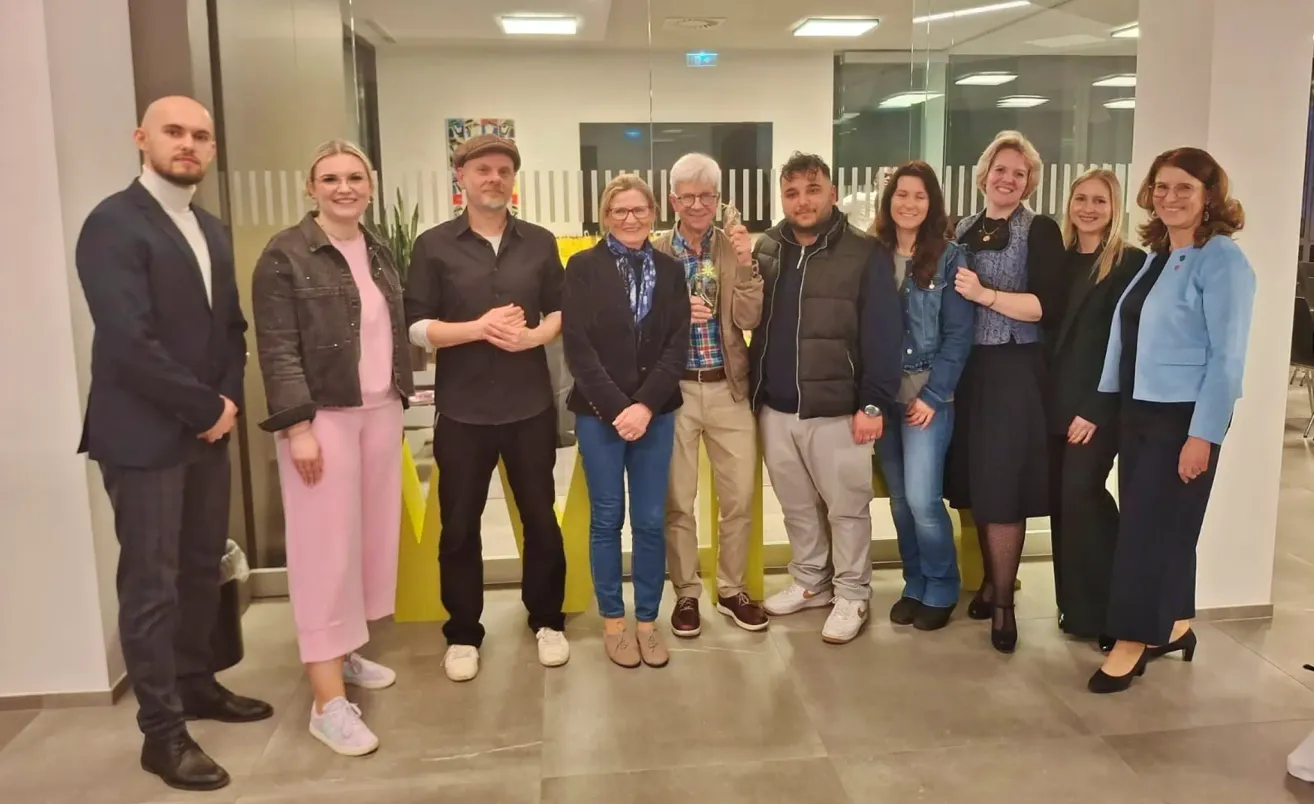 A group of people are posing for a photo in a modern office. The man in the middle holds a bottle. The people are dressed in various outfits, and there are yellow structures behind them.