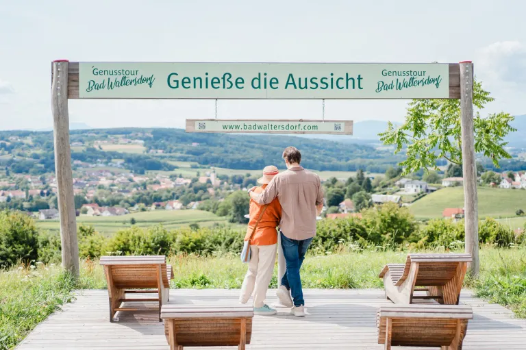 A couple stands on a wooden deck, looking at a scenic view of a town with hills and mountains in the distance. A sign above them reads, Genieße die Aussicht.
