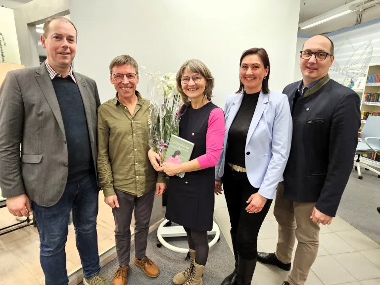 Fünf Personen posieren lächelnd für ein Foto. Eine Frau in der Mitte hält ein Buch und einen Blumenstrauß. Die Frau rechts trägt eine hellblaue Jacke.