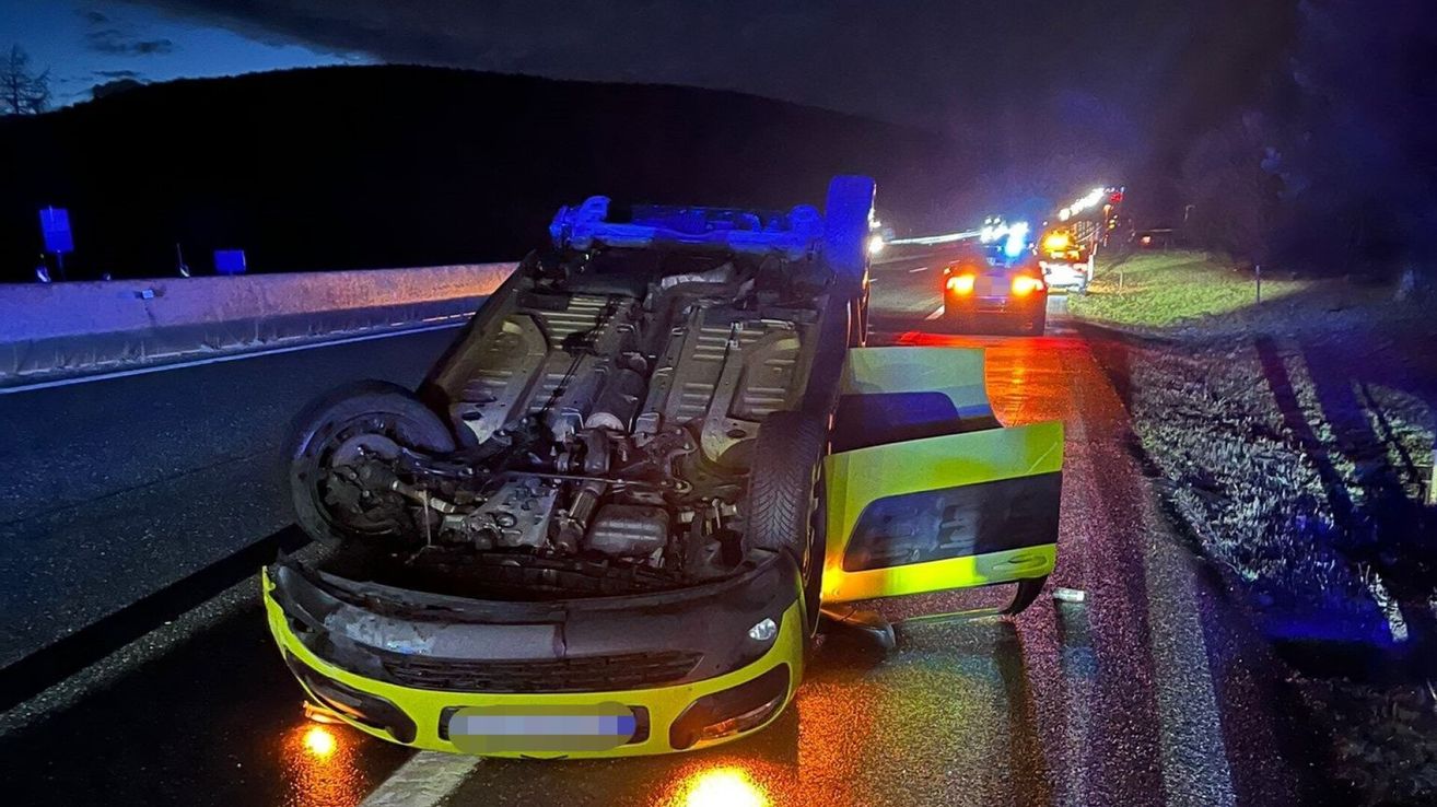 An overturned sports car with its engine exposed lies on a highway at night, with emergency lights illuminating the scene.