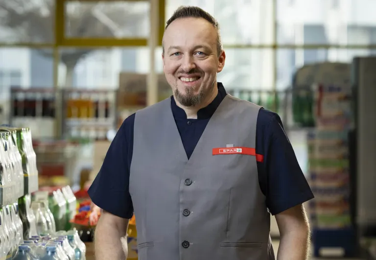 Ein Mann in einer grauen Weste mit einem roten Schild, das 'SPAR' steht, lächelt und steht in einem Geschäft.