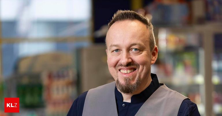 Ein Mann mit Bart lächelt in die Kamera, trägt ein blaues Hemd und eine graue Weste und steht vor einem verschwommenen Hintergrund.