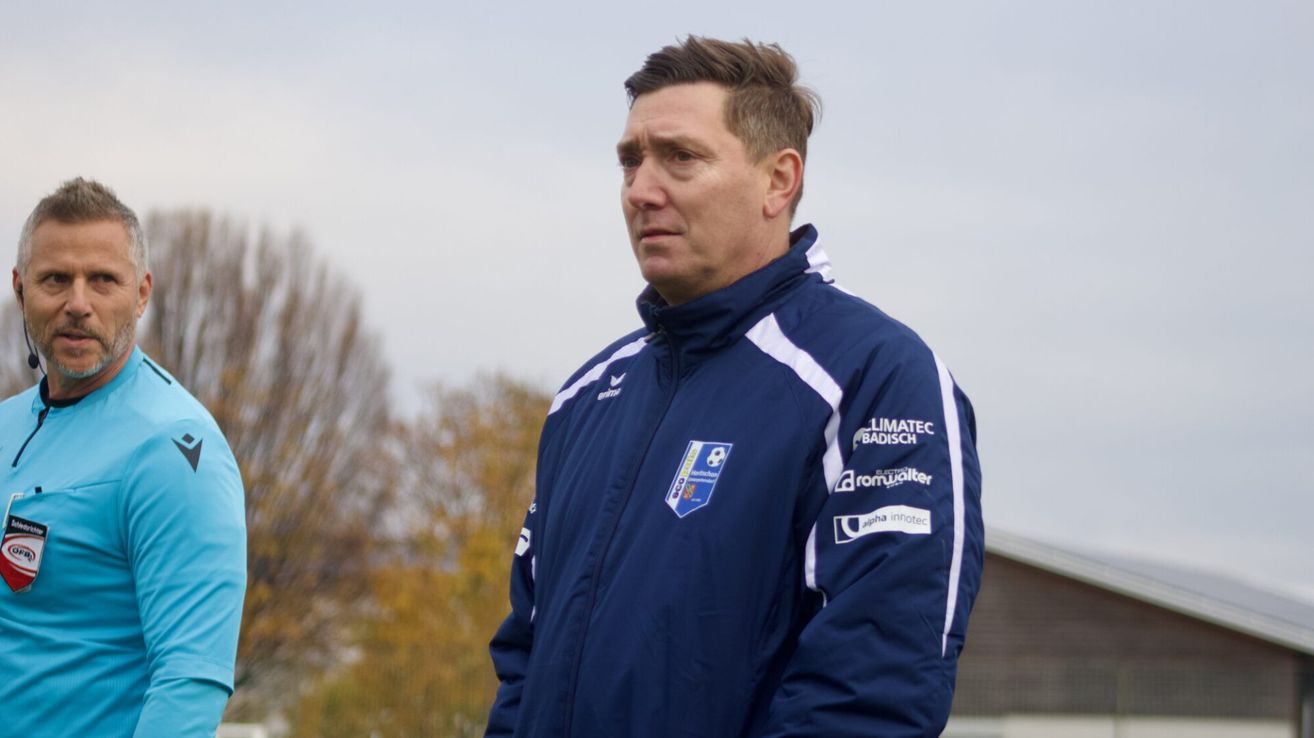 Ein Mann in einem blauen Jackett mit weißen Streifen steht draußen. Das Jackett hat mehrere Logos, darunter 'Limatec Badisch', 'romwalter' und 'alpha innotec'. Der Hintergrund zeigt Bäume.