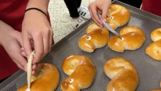 Mehrere Personen formen Brezeln in Form von Fischen auf einem Backblech. Die Brezeln haben mit einer weißen Substanz hinzugefügte Augen. Die Bäcker tragen rote Schürzen.