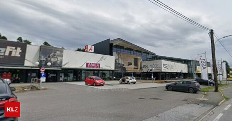 Ein Einkaufskomplex mit drei Geschäften, darunter Amica und Asslmbel. Ein rotes Auto und ein weißes Auto sind vor dem Gebäude geparkt. Eine Person steht neben dem weißen Auto. Ein schwarzes Auto fährt auf der Straße.