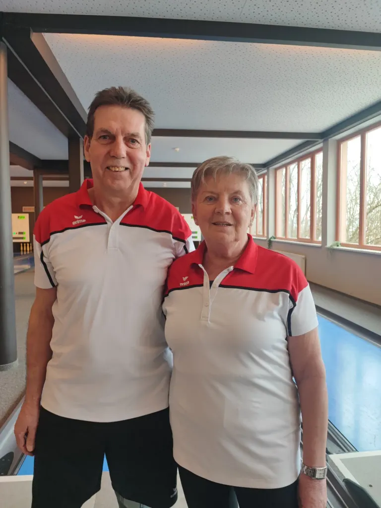 Ein Mann und eine Frau lächeln und stehen zusammen in einer Bowlingbahn. Sie tragen beide weiße und rote Poloshirts.
