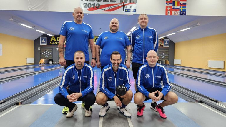 Fünf Männer in blauen Trikots mit weißen Streifen posieren für ein Foto in einer Bowlingbahn. Sie lächeln und scheinen für ein Foto zu posieren. Hinter ihnen ist ein Banner mit dem Text '2010' und einem Logo.