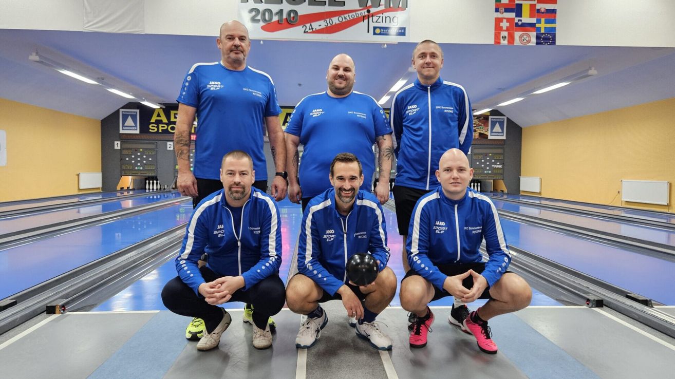 Fünf Männer in blauen Trikots mit weißen Streifen posieren für ein Foto in einer Bowlingbahn. Sie lächeln und scheinen für ein Foto zu posieren. Hinter ihnen ist ein Banner mit dem Text '2010' und einem Logo.