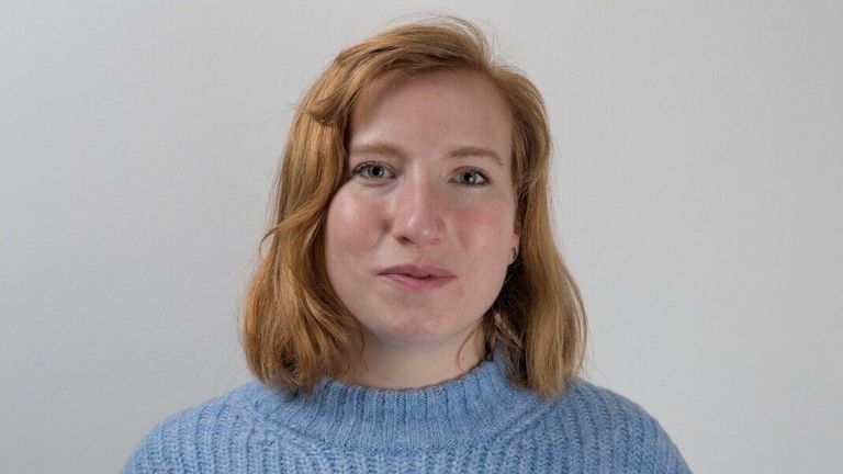 Eine Frau mit kurzen blonden Haaren und einem hellblauen Rollkragenpullover posiert vor einem weißen Hintergrund.