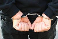 Eine Person in schwarzer Kleidung ist mit silbernen Ketten an den Handgelenken gefesselt und hält ihre Hände vor sich zusammen.