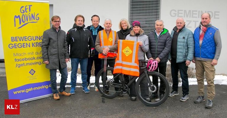Eine Gruppe von Menschen steht draußen in Winterkleidung. Einer hält eine Flagge und ein anderer ein Fahrrad mit Helm. Hinter ihnen ist ein gelber Banner mit Text.
