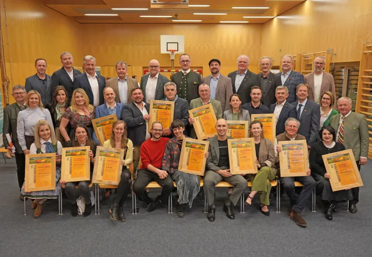 Eine Gruppe von Personen, sowohl Männer als auch Frauen, sind in einem Raum versammelt. Sie sitzen auf Stühlen und stehen. Sie halten alle Urkunden in den Händen. Sie alle lächeln. Sie tragen formelle Kleidung. Sie posieren für ein Gruppenfoto. Der Raum hat Holzwände und einen Basketballkorb.