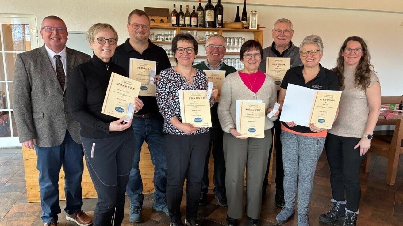 Eine Gruppe lächelnder Menschen posiert für ein Foto und hält Urkunden in der Hand. Sie stehen in einem Raum mit Regalen, die mit Flaschen und Gläsern gefüllt sind, im Hintergrund.