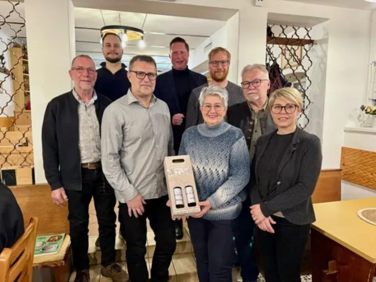 Eine Gruppe von Erwachsenen, einschließlich Männer und Frauen, posiert für ein Foto, während sie eine Box halten. Sie stehen auf den Treppen mit einer warmen, gemütlichen Atmosphäre. Sie tragen alle eine Brille und lächeln. Dahinter befindet sich eine weiße Wand mit einer Lampe, die von der Decke hängt.