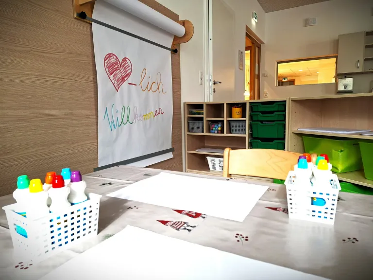 Ein Klassenzimmer mit einem Schreibtisch und Stühlen. Ein Roll-up-Banner mit einem Herz und dem Text 'Willkommen' hängt an der Wand. Ein Regal mit Aufbewahrungsboxen und einer Tür, die in einen anderen Raum führt.