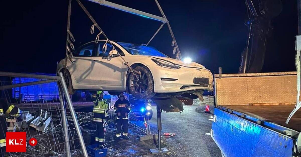 Ein weißer Tesla Model 3 wird bei Nacht von einem Kran gehoben. Zwei Feuerwehrleute stehen in der Nähe, einer in einer gelben Weste. Die Front des Autos ist beschädigt.