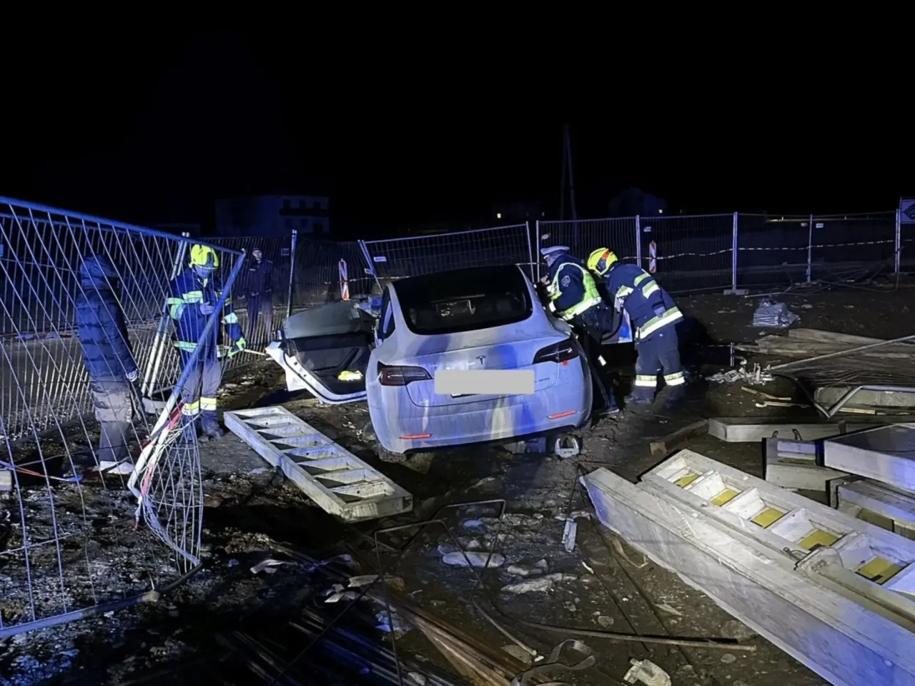 Ein Tesla Model 3 ist nachts beschädigt, und Rettungskräfte kümmern sich um das Fahrzeug. Das Auto ist gekippt, und der vordere Stoßfänger ist beschädigt. Die Arbeiter tragen Schutzausrüstung.