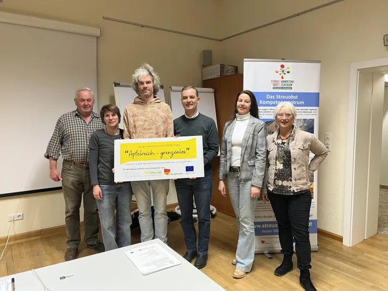 Fünf Erwachsene stehen lächelnd in einem Raum und halten ein gelbes Schild mit der Aufschrift Apfelreich-grenzenlos. Ein Banner mit dem Text Das Streuobst Kompetenzzentrum steht hinter ihnen.