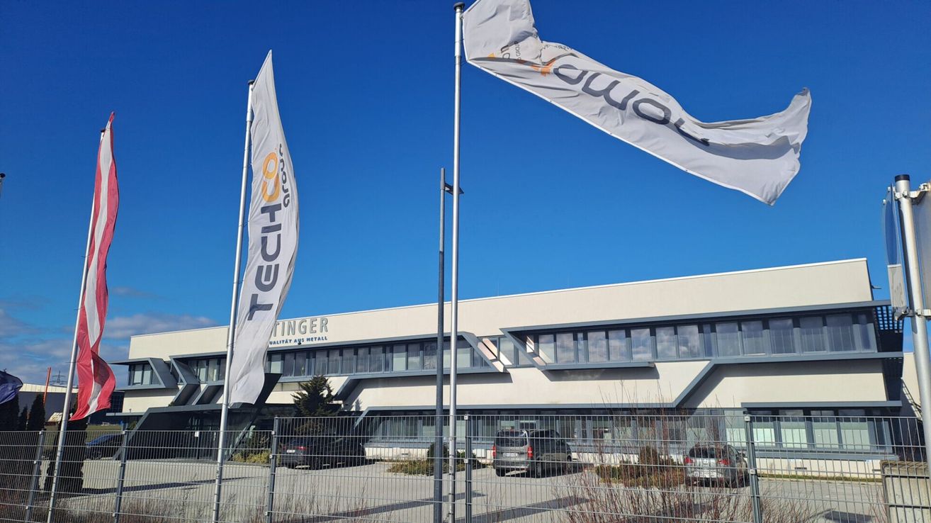 Ein modernes Gebäude mit weißen Fahnen davor, eine mit dem Wort 'Techco' und die andere mit 'Finger'. Autos sind vor dem Gebäude unter einem klaren Himmel geparkt.