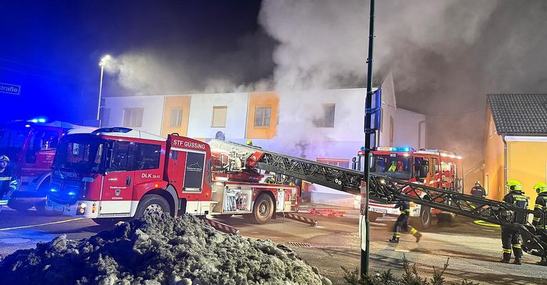 Feuerwehrleute reagieren auf einen Brand in einem Gebäude mit orangefarbenen Akzenten. Zwei Feuerwehrwagen sind am Einsatzort, einer mit seiner Leiter zum Gebäude ausgestreckt. Rauch ist sichtbar.