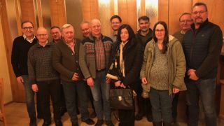 Eine Gruppe von Personen posiert für ein Foto in einem Raum mit Holzwänden. Sie sind in Winterkleidung gekleidet. Eine Frau in der Mitte hält eine schwarze Tasche.