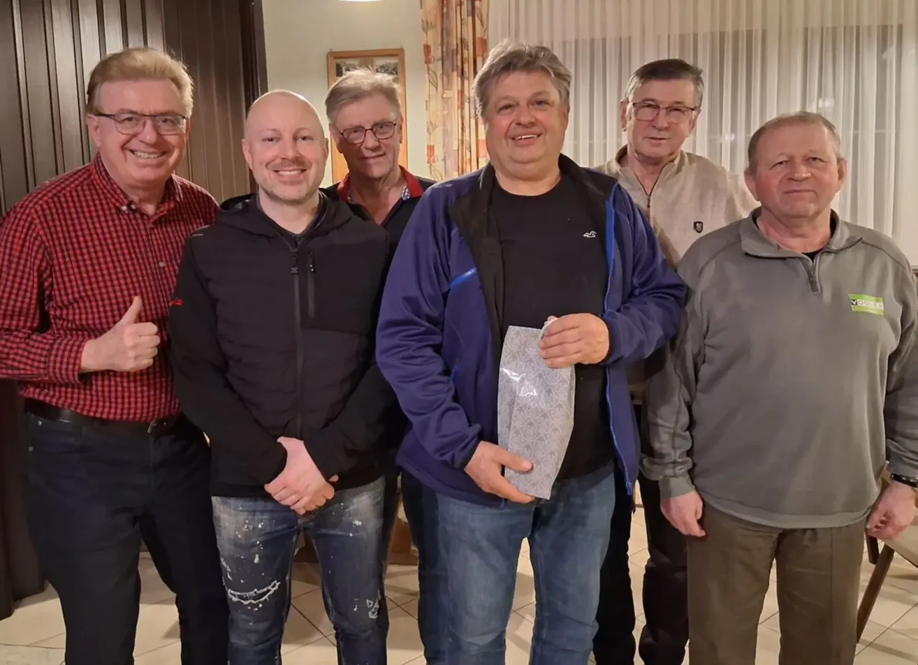 Fünf Männer stehen zusammen in einem Raum, lächeln und posieren für ein Foto. Ein Mann in der Mitte hält eine Auszeichnung und lächelt. Der Mann auf der linken Seite trägt ein rot kariertes Hemd.