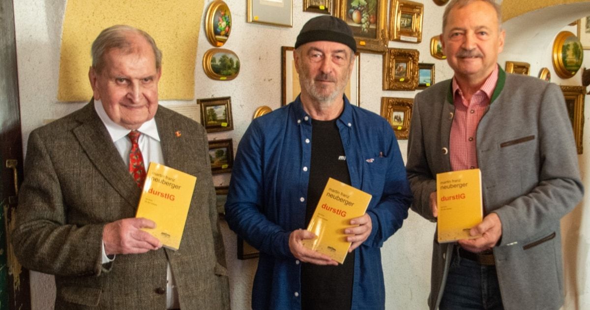 Drei Männer stehen zusammen, lächeln und halten gelbe Bücher mit dem Titel 'DURSTI'G'. Der Mann auf der linken Seite trägt einen Anzug und eine Krawatte. Der Mann in der Mitte trägt ein blaues Hemd und einen schwarzen Hut. Der Mann auf der rechten Seite trägt ein rosa Hemd. Hinter ihnen hängen mehrere gerahmte Bilder an der Wand.