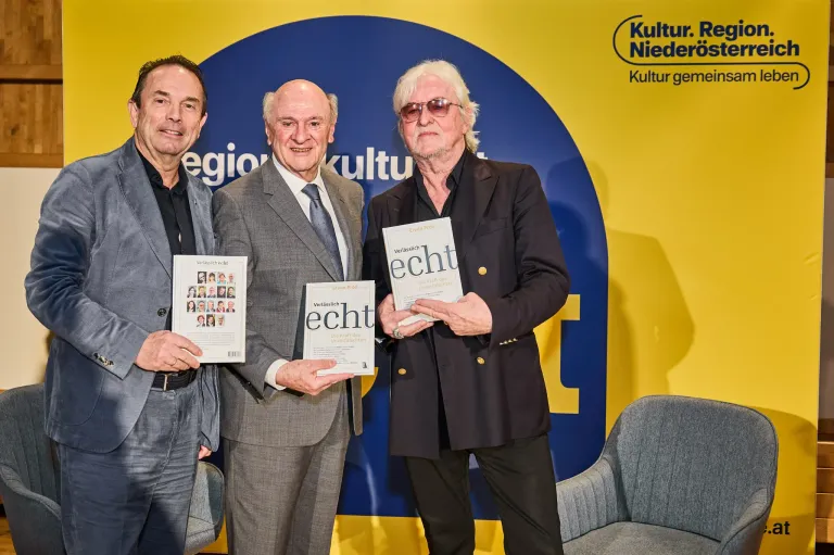 Drei Männer stehen vor einem gelben und blauen Hintergrund mit Logos und halten Bücher. Der Mann auf der linken Seite trägt einen Anzug und hält ein Buch mit einem Foto von Menschen. Der Mann in der Mitte trägt einen Anzug und eine Brille und hält ein Buch mit dem Titel 'Veritaslich'. Der Mann auf der rechten Seite trägt einen schwarzen Anzug und hält ein Buch mit dem Titel 'Echt'.