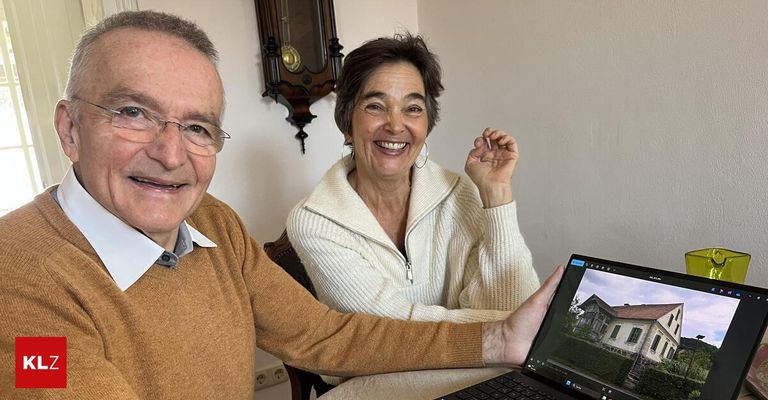 An older couple sitting together, smiling, and looking at a photo of a house on a laptop. The man holds the laptop while the woman gestures. A wall clock and power outlet are in the background.