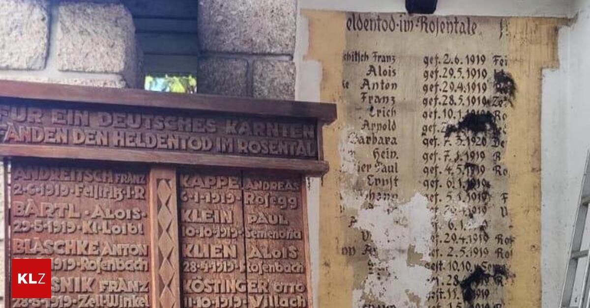 Eine Gedenktafel für gefallene Soldaten ist an einer Steinmauer angebracht. Namen und Geburts- und Todesdaten sind darauf eingemeißelt. Ein altes, zerrissenes Dokument mit handgeschriebenem Text ist daneben platziert.