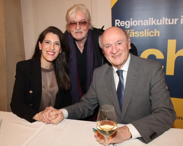 Drei Personen sitzen gemeinsam an einem Tisch, lächeln und halten Händchen. Der Mann auf der rechten Seite hält ein Weinglas, und sie sind alle formell gekleidet. Hinter ihnen befindet sich ein Banner mit Text in einer fremden Sprache.