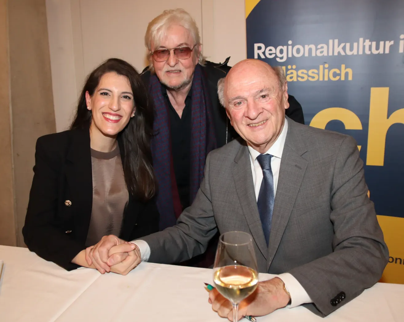 Three individuals sit together at a table, smiling and holding hands. The man on the right holds a wine glass, and they are all dressed formally. Behind them is a banner with text in a foreign language.