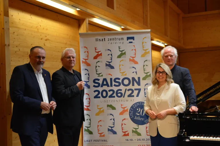Vier Personen stehen vor einem Banner. Es zeigt mehrere Porträts und den Text 'LISZT ZENTRIUM Raiding SAISON 2026/27'. Die Frau lächelt und hält ein Stück Papier in der Hand.