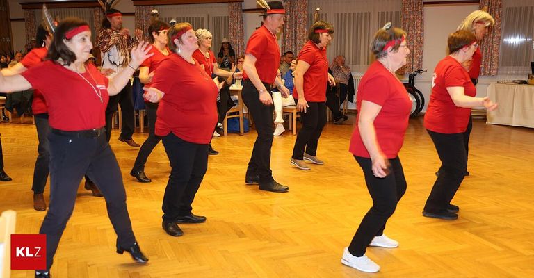 Eine Gruppe von Menschen in roten Shirts und schwarzen Hosen tanzt auf einem Holzboden. Einige tragen Stirnbänder. Andere beobachten vom Stuhl aus. Ein Tisch hält einen Becher.