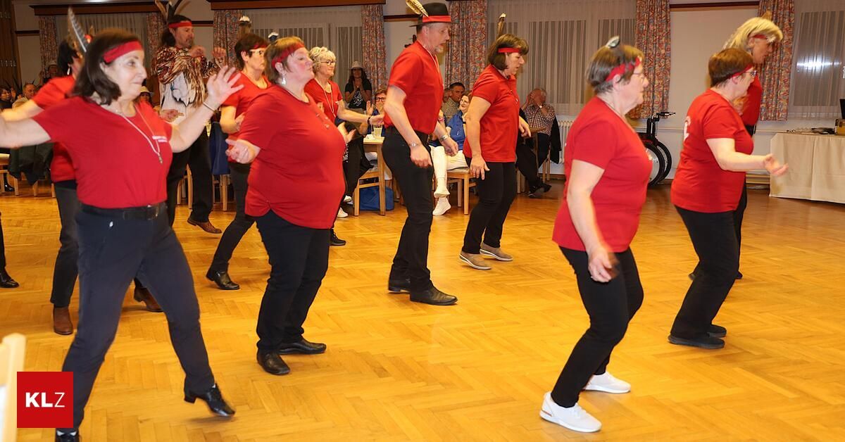 Eine Gruppe von Menschen in roten Shirts und schwarzen Hosen tanzt auf einem Holzboden. Einige tragen Stirnbänder. Andere beobachten vom Stuhl aus. Ein Tisch hält einen Becher.