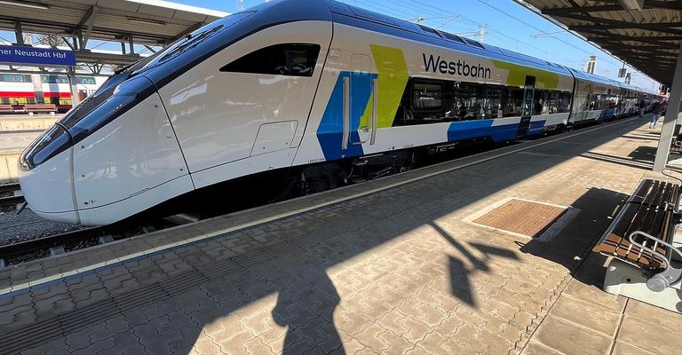 A Westbahn train stands at a platform under a bright sky, with a person's shadow cast on the ground.