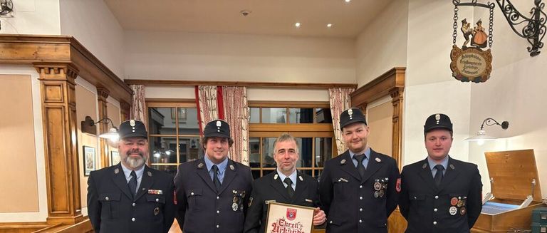 Fünf Feuerwehrleute in Uniform stehen zusammen drinnen, einer hält ein gerahmtes Zertifikat. Hinter ihnen befindet sich ein Raum mit einer Holzdecke, einem dekorativen Schild und einem Fenster mit roten Vorhängen.